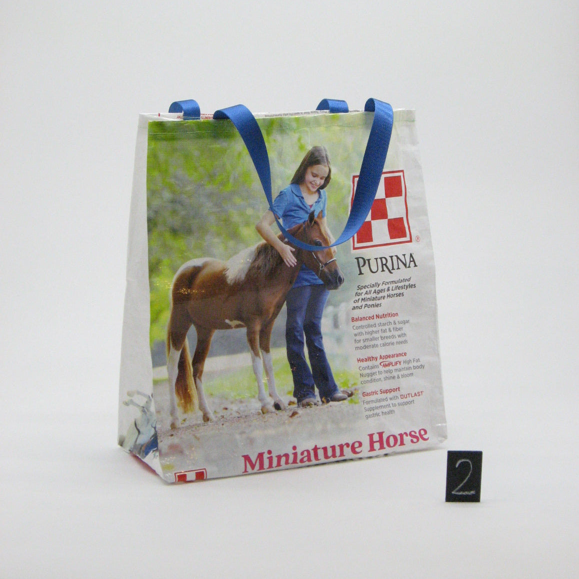 A white Purina Miniature Horse promotional tote bag with blue handles, featuring a printed photo of a woman leading a brown horse, displayed on a white surface.