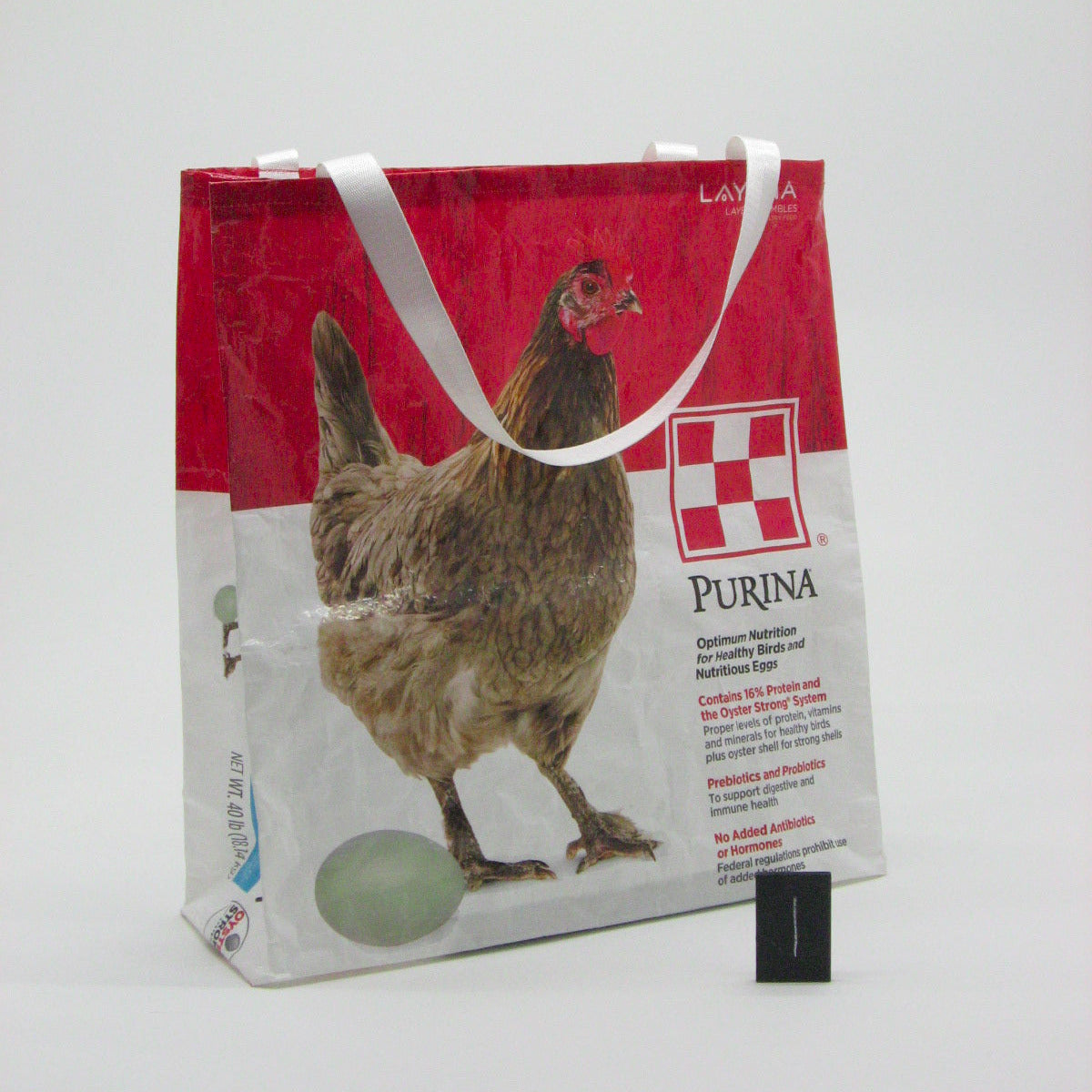 A custom printed recycled paper tote bag with white handles, featuring a red top section, a white bottom section, a brown chicken graphic, and the Purina logo. The bag is photographed on a plain white studio background with a small black scale card in front.