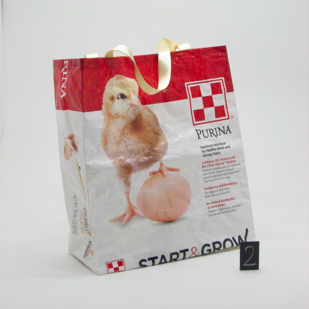 A Purina Start & Grow paper shopping bag with gold handles, featuring a printed image of a baby chick and a peach egg on a white and red background, photographed on a white studio background.