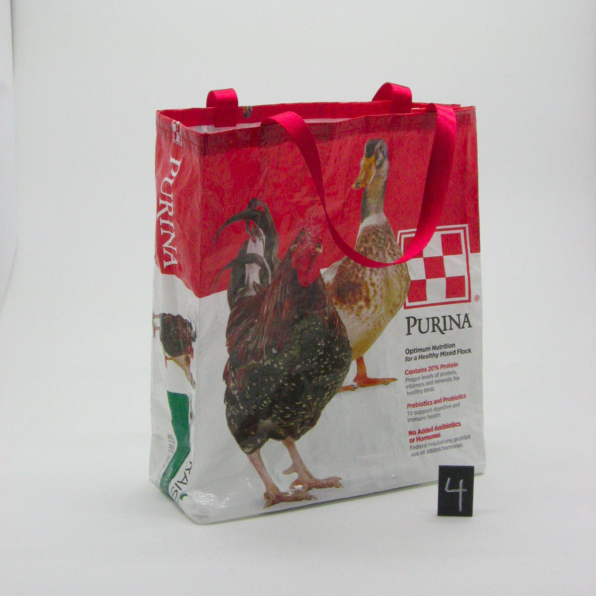 A reusable Purina branded bag with red handles, featuring printed images of chickens and a red-and-white logo panel, photographed on a light gray studio background.