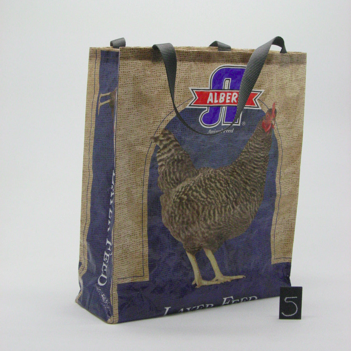 A reusable jute tote bag with black handles, featuring a blue and tan design with a rooster illustration and the 'Alber' logo. A small black card with the number 5 is placed next to it on a light gray background.