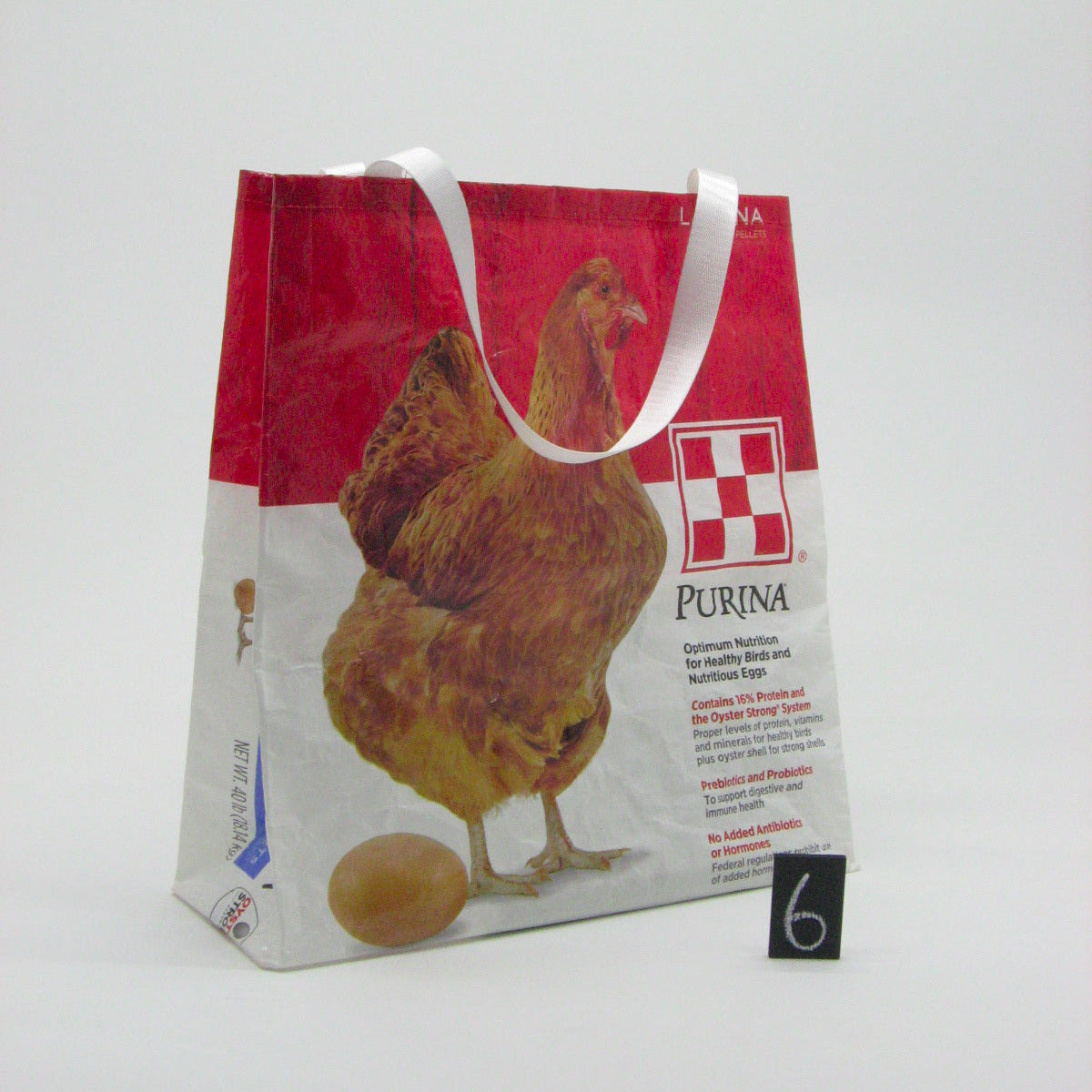 A reusable paper shopping bag with white handles, featuring a red top section, a printed brown chicken and egg, and the Purina logo on a white studio background.