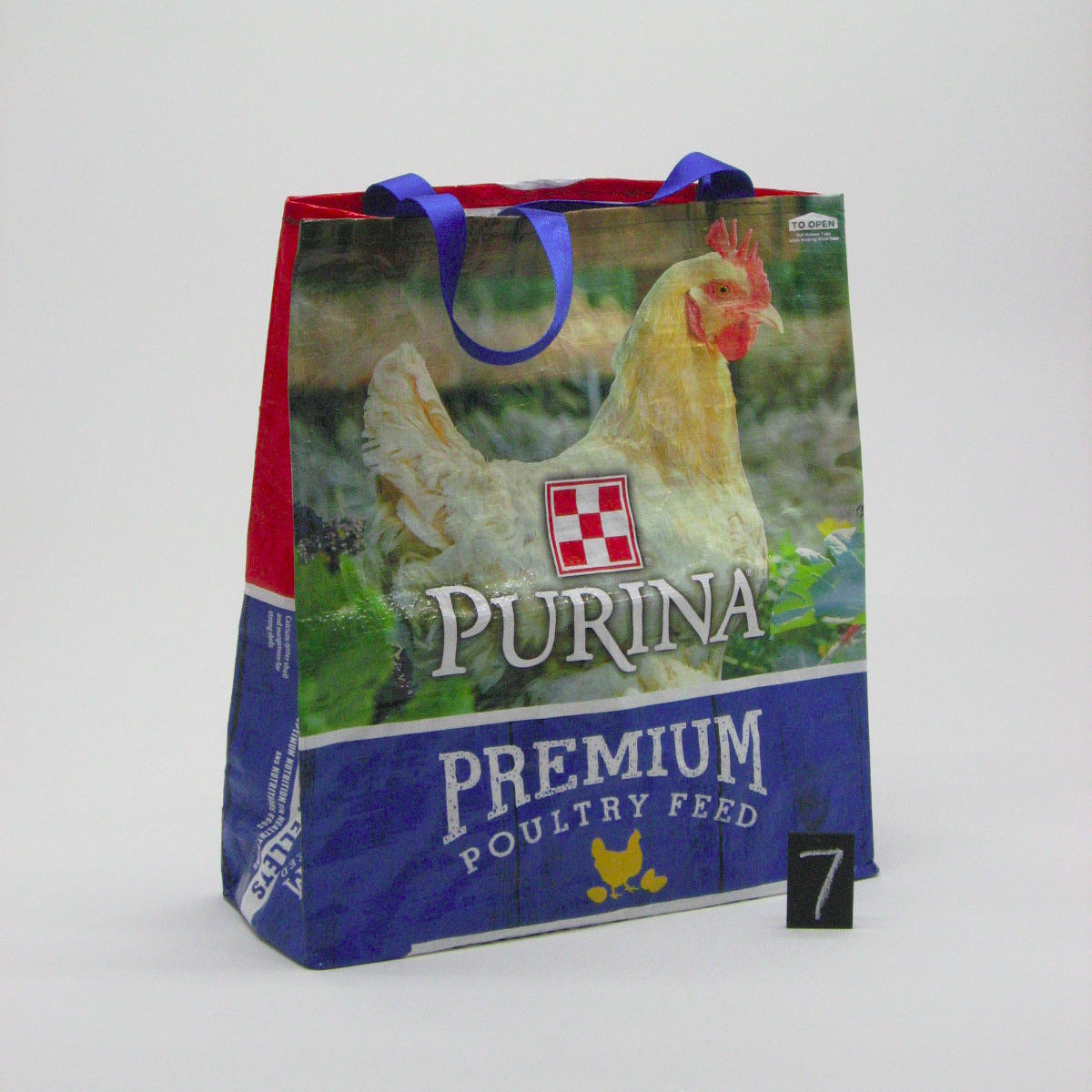 A Purina Premium Poultry Feed reusable shopping bag with blue handles, featuring a printed image of a chicken and the Purina brand logo, set against a plain light gray background.