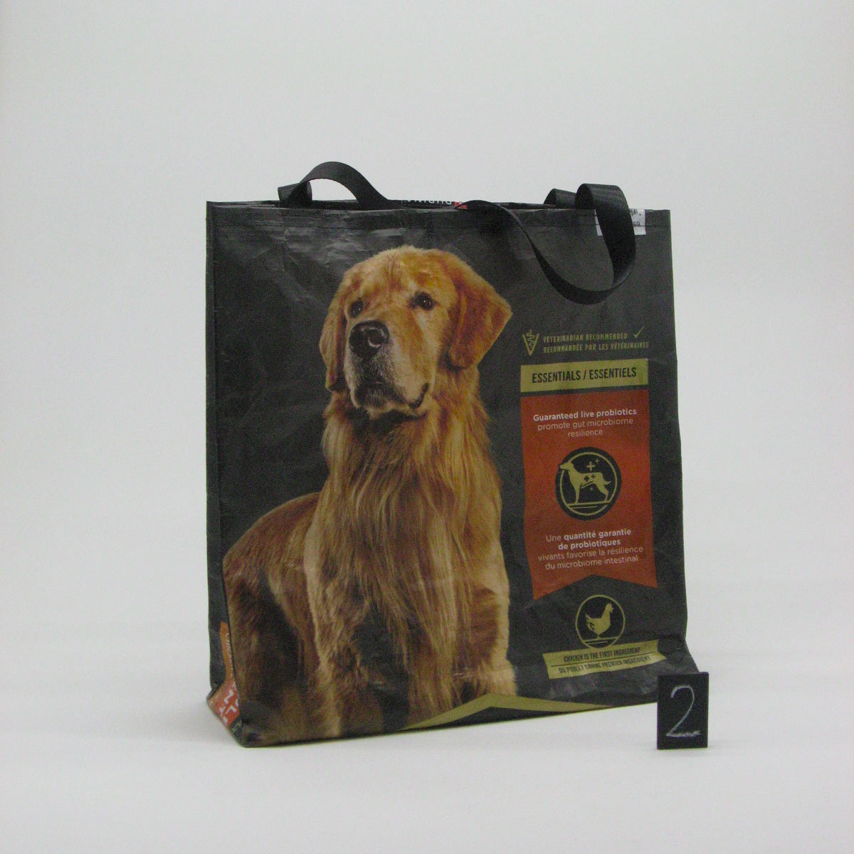 A black reusable tote bag featuring a large printed photo of a golden retriever dog and a vertical orange and gold label panel, photographed on a light gray studio background with a small black '2' card in front.