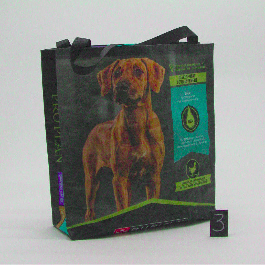A reusable black shopping tote bag with a printed image of a brown dog and product information panels, standing on a white studio background next to a small black number 3 card.