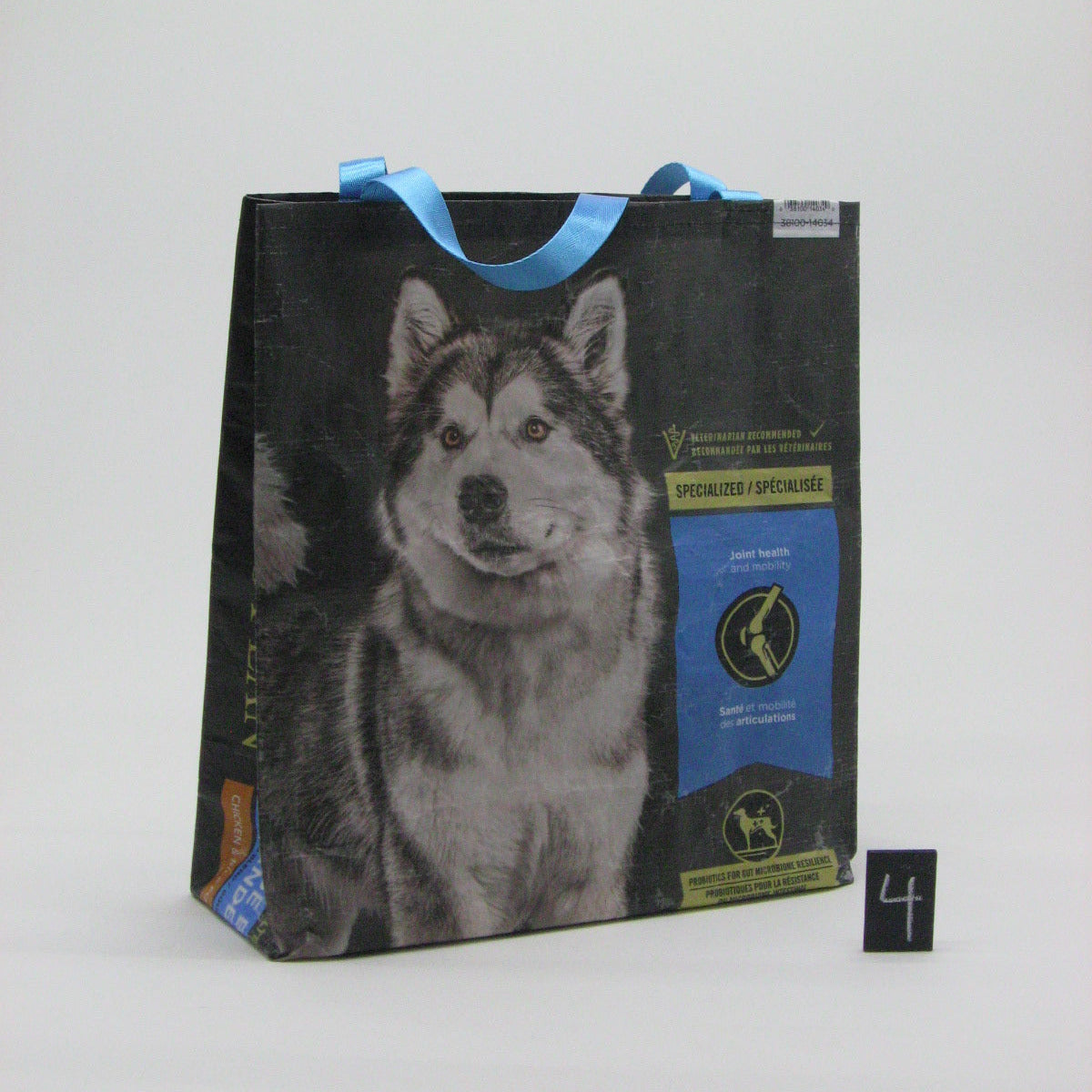 A reusable paper shopping bag with blue handles, featuring a large photo of a husky dog on the front. The bag has a black background with a blue and gold label on the right side, photographed on a white studio background with a small black number '4' card next to it.