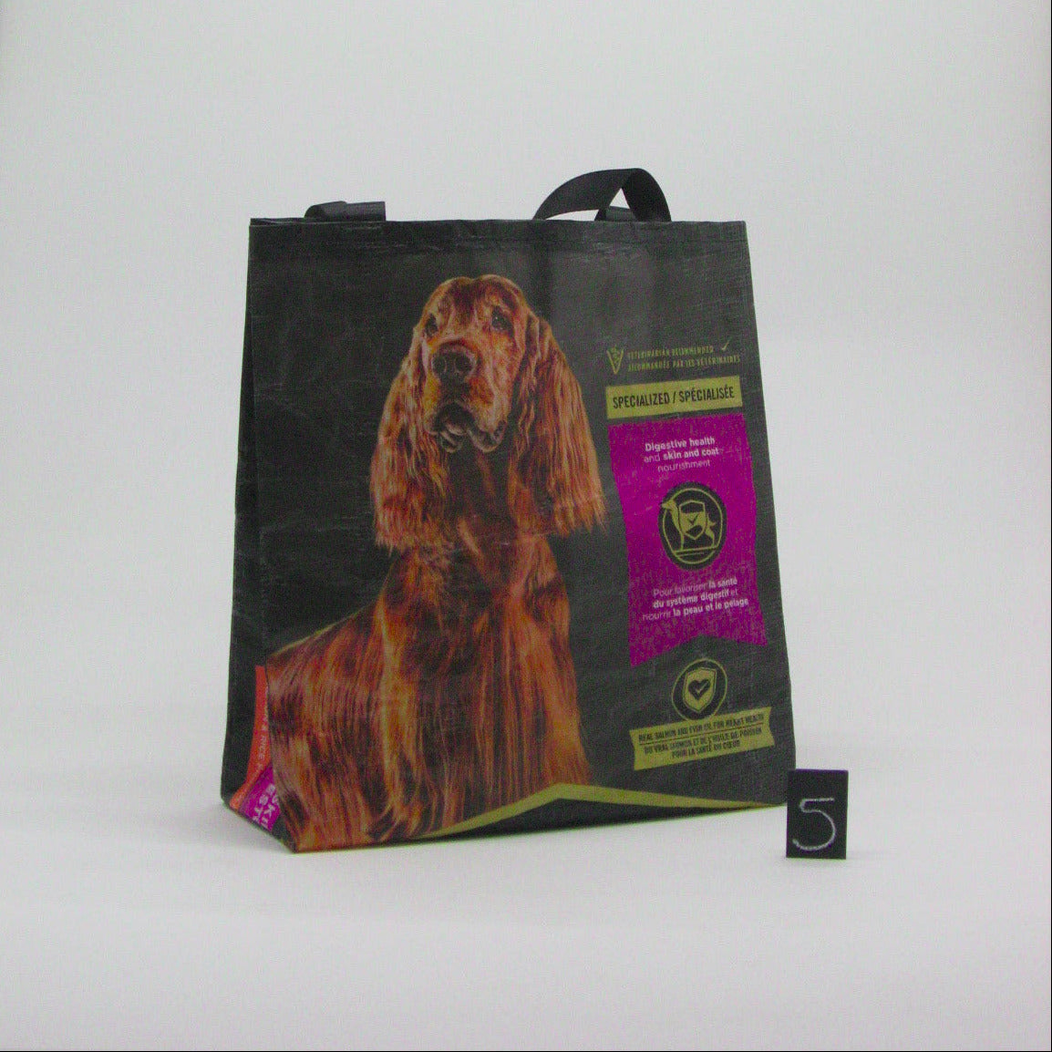 A black reusable shopping tote bag featuring a large print of a brown long-haired dog and a purple and gold dog food label, standing on a white studio background next to a small black scale card.